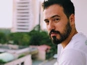 A serious-looking man with a beard stands on a balcony with blurred city buildings in the background.